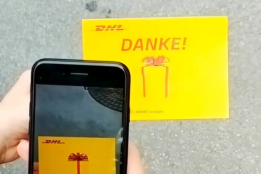 A phone over a yellow DHL DANKE postcard on the pavement; on the screen the drawn gift gets a red bow