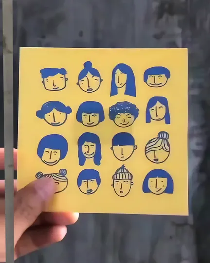A yellow print of sixteen ink-drawn faces held up in a hand; through the phone the same faces change and come to life inside a blue frame